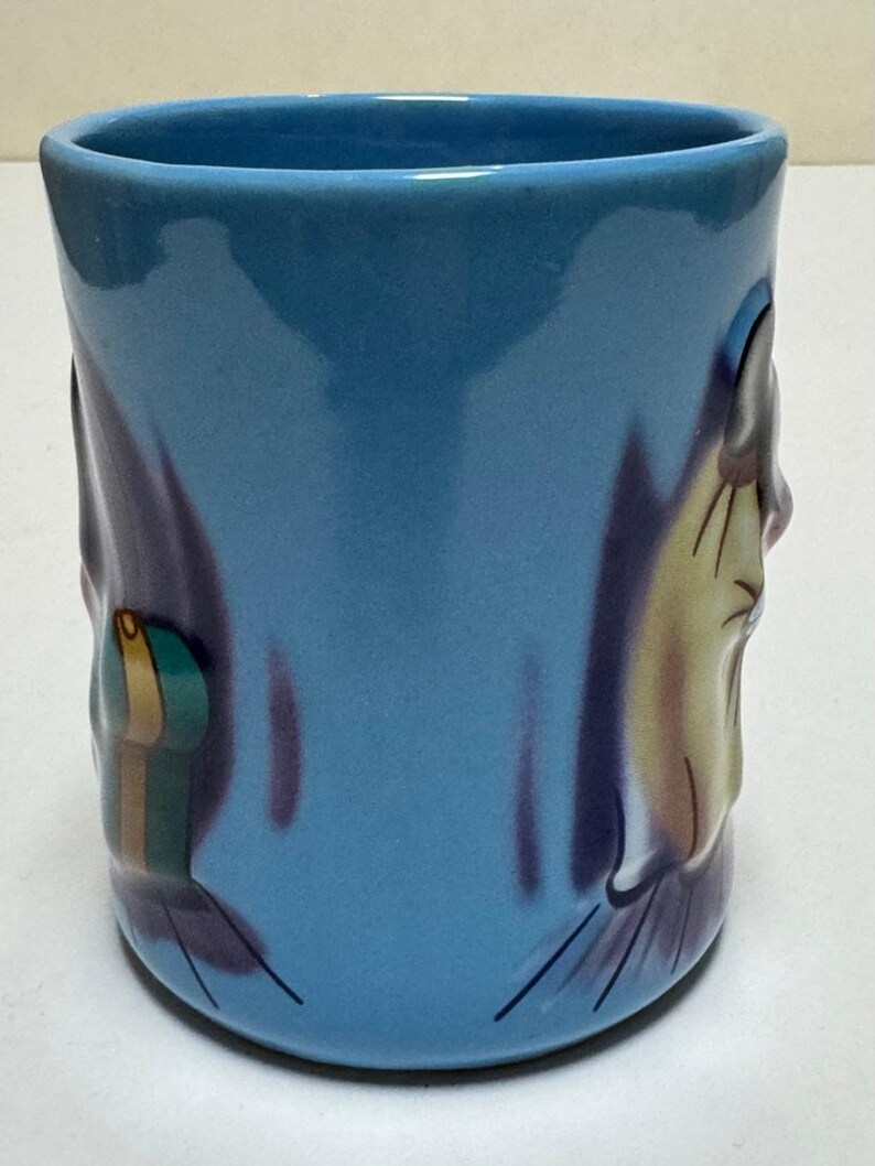 May include: A blue ceramic mug with a cartoon-style design. The mug features a light blue exterior with a darker blue and purple design on the sides. The design includes a cartoon face and a small, colorful object.