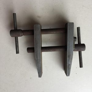 以下が含まれることがあります： 2本の平行なネジ棒と2本の垂直なバーを備えたダークグレーの金属製ハンドクランプ。ネジ棒の端には2つの垂直なハンドルが付いており、バーの間の距離を手動で調整できるようになっています。