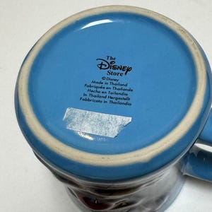 May include: A blue ceramic mug with a white rim and a handle. The bottom of the mug features the text "The Disney Store" and "Made in Thailand". The mug has a dark brown design on the exterior.