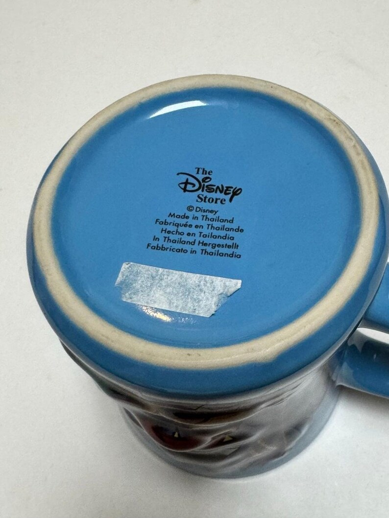 May include: A blue ceramic mug with a white rim and a handle. The bottom of the mug features the text "The Disney Store" and "Made in Thailand". The mug has a dark brown design on the exterior.