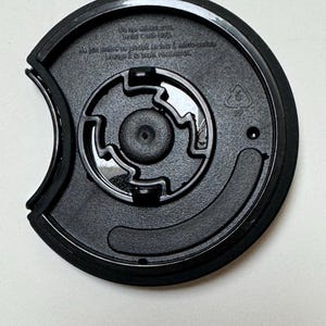 May include: Black plastic cup lid with a circular design. The lid has a recessed center with a smaller circular piece in the middle. The lid has text that reads "Do not microwave. Hand wash only."