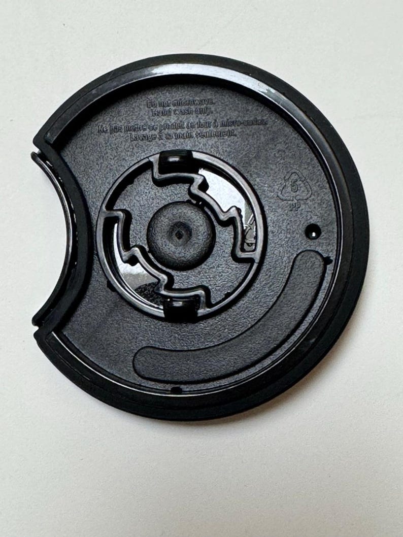 May include: Black plastic cup lid with a circular design. The lid has a recessed center with a smaller circular piece in the middle. The lid has text that reads "Do not microwave. Hand wash only."