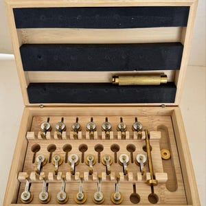 Può includere: Una scatola di legno con coperchio incernierato, contenente una serie di piccoli utensili di precisione. Gli utensili sono disposti in file e sono in metallo e legno. La scatola ha una chiusura dorata e una maniglia.