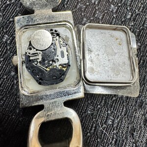 May include: A silver-toned wristwatch with the back open, revealing the internal mechanism and a small battery. The watch has a rectangular face and a leather strap. The watch face is open, showing the internal components.