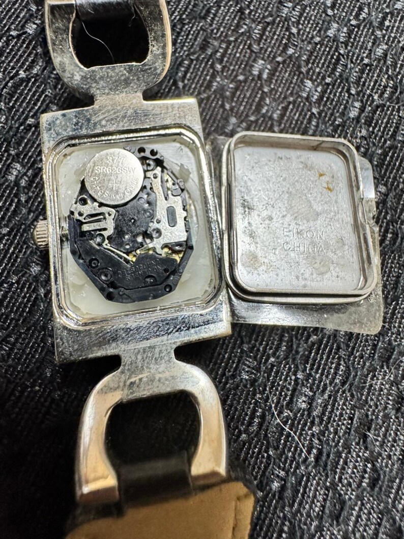 May include: A silver-toned wristwatch with the back open, revealing the internal mechanism and a small battery. The watch has a rectangular face and a leather strap. The watch face is open, showing the internal components.