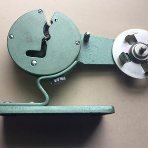 May include: A sage green metal tape dispenser featuring a circular housing with a central slot and a side-mounted silver metal spool holder with four prongs. The device is mounted on a flat, rectangular base with a curved support arm and a tension knob.