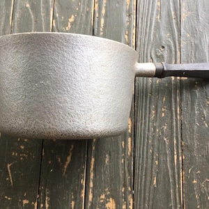 Puede incluir: Un cazo de hierro fundido gris con un acabado mate y texturizado y un mango recto de color gris oscuro que se extiende desde el lateral. El mango cuenta con un pequeño lazo de metal en el extremo para colgarlo. La olla tiene una forma cilíndrica.