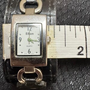 May include: A silver-toned Eikon quartz watch with a white face, black numerals, and green hands. The watch is attached to a black leather cuff-style band. The watch face is approximately 1 inch wide.