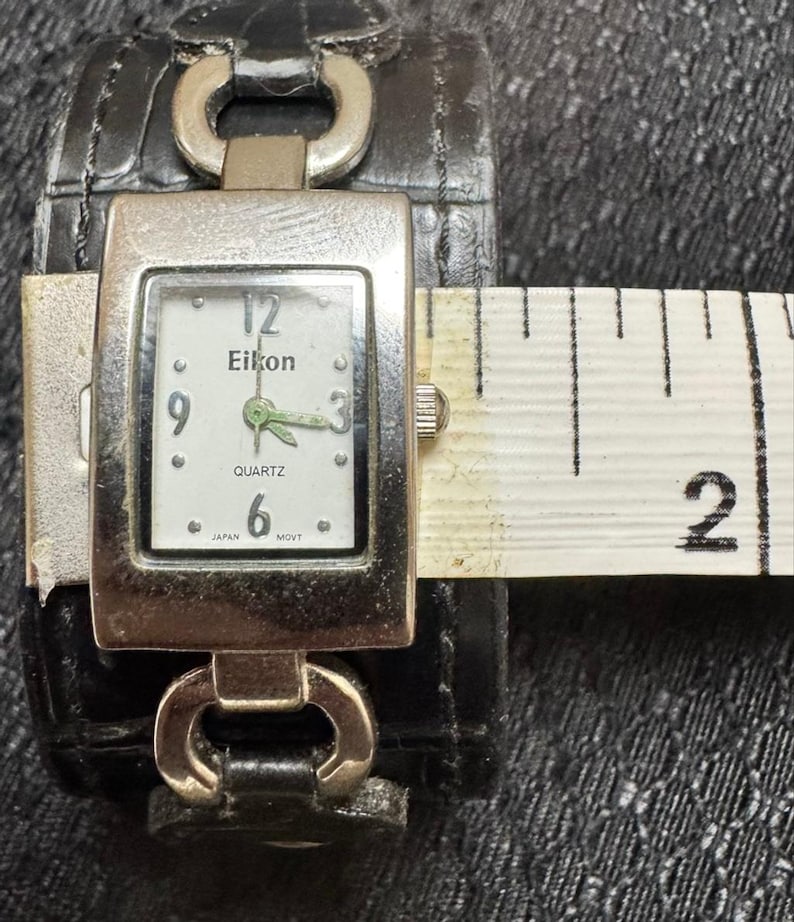 May include: A silver-toned Eikon quartz watch with a white face, black numerals, and green hands. The watch is attached to a black leather cuff-style band. The watch face is approximately 1 inch wide.