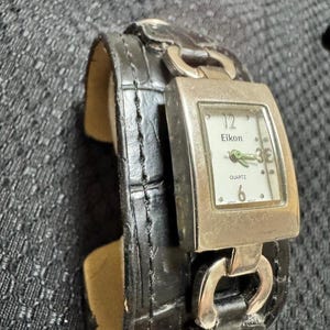 May include: A black leather cuff watch with a silver-toned rectangular watch face. The watch face displays the brand name "Eikon" and the word "Quartz". The watch band has a crocodile skin pattern.