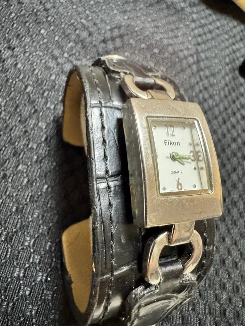 May include: A black leather cuff watch with a silver-toned rectangular watch face. The watch face displays the brand name "Eikon" and the word "Quartz". The watch band has a crocodile skin pattern.