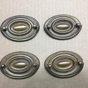 May include: Four oval-shaped brass drawer pulls with a dark, aged patina. Each pull features a central raised oval element surrounded by concentric rings and a hinged, curved handle attached to two small ball-shaped mounting points on the sides.