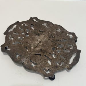 May include: An oval-shaped silver-toned metal trivet featuring an intricate openwork scroll and floral pattern. The piece is composed of two symmetrical halves joined together and is supported by small, dark, rounded feet at the base.