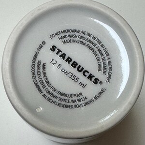May include: Bottom of a white Starbucks coffee mug. The text includes "STARBUCKS" and "12 fl oz/355 ml". Additional text indicates the mug is not microwave safe and should be hand washed. Made in China.