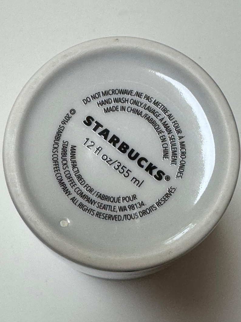 May include: Bottom of a white Starbucks coffee mug. The text includes "STARBUCKS" and "12 fl oz/355 ml". Additional text indicates the mug is not microwave safe and should be hand washed. Made in China.