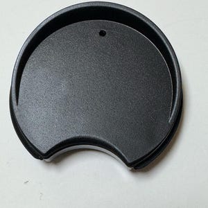 May include: A black, semi-circular lid with a small hole, likely for a travel mug or cup. The lid has a textured surface and rounded edges, with a design reminiscent of mouse ears.