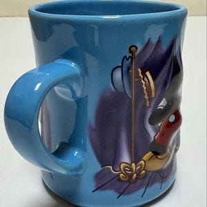 May include: A light blue ceramic mug with a curved handle. The mug features a cartoon illustration of a character in purple, black, and red. The handle and interior of the mug are the same light blue color.