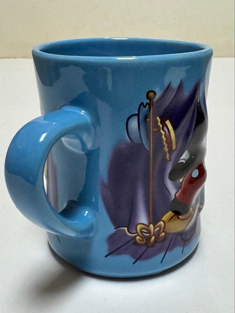 May include: A light blue ceramic mug with a curved handle. The mug features a cartoon illustration of a character in purple, black, and red. The handle and interior of the mug are the same light blue color.