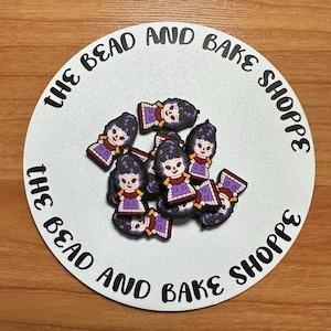 May include: A collection of doll-shaped charms with black hair, purple dresses, and red accents. The charms are arranged on a white circular surface with the text "THE BEAD AND BAKE SHOPPE" in a black, curved font.