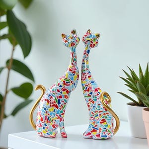 Peut inclure: Deux figurines décoratives de chats. Les chats ont de longs cous et queues, avec un motif de mosaïque de formes géométriques colorées. Les oreilles et les queues sont dorées. Les chats sont posés face à face.