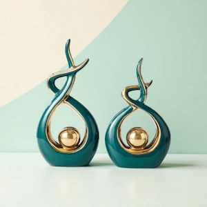 Könnte beinhalten: Zwei teal- und goldfarbene Dekorationsskulpturen. Jede Skulptur hat eine tropfenförmige Form mit einer goldenen Kugel in der Mitte und goldenen Akzenten. Die Skulpturen haben ein modernes, abstraktes Design, vor hellgrünem und weißem Hintergrund.