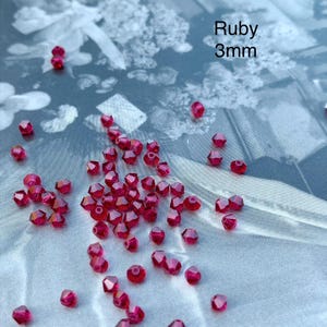 Peut inclure: Une collection de petites perles facettées de couleur rubis dispersées sur la surface. Chaque perle mesure 3 mm de diamètre. Le texte Ruby 3mm est clairement visible dans le coin supérieur droit de l'image, indiquant la couleur et la taille.