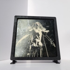 May include: A black framed art piece featuring a stylized illustration of a figure with outstretched arms. The artwork is primarily black and white with red accents, set against a textured background. The frame has a square shape and is supported by two small feet.