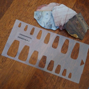 May include: A clear plastic template with the text "PEDESTALS 1" and multiple oval-shaped cutouts of varying sizes. The template is on a wooden surface with three pieces of colorful, patterned stone.