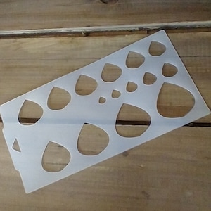 May include: A white plastic stencil with multiple teardrop shapes of varying sizes. The stencil is designed for crafting and creating projects.