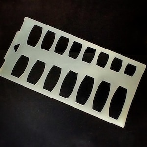 May include: A translucent, rectangular stencil with a series of oval cutouts. The stencil is made of a light-colored material and is set against a dark background. The cutouts are evenly spaced.