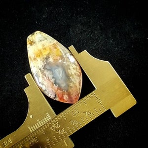 May include: A close-up of a colorful agate cabochon, measuring approximately 2.5 inches in length, being measured with a ruler. The agate features a variety of colors, including red, orange, yellow, white, and gray.