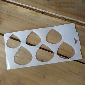 May include: A white plastic stencil with six teardrop-shaped cutouts. The stencil is designed for crafting and creating projects.