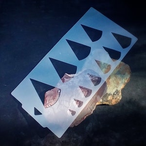 May include: A clear plastic stencil with multiple triangle shapes cut out. The stencil is lying on a brown and green rock.