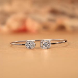 Könnte beinhalten: Ein silberfarbenes Armband mit zwei diamantbesetzten Fassungen. Eine Fassung ist rund, die andere quadratisch. Das Armband wird auf einer hellbraunen Oberfläche mit unscharfem Hintergrund präsentiert.