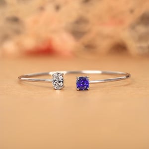 Könnte beinhalten: Ein filigranes Silberarmband mit einem funkelnden, rund geschliffenen Diamanten und einem quadratischen, tiefblauen Edelstein. Das Armband hat ein minimalistisches Design, wobei die Edelsteine an beiden Enden des schmalen Bandes angebracht sind.