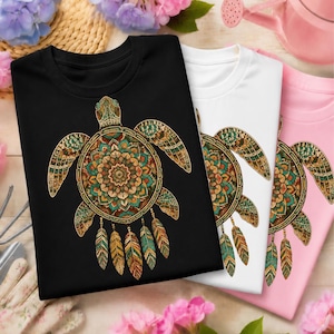 May include: A black, a white, and a light pink t-shirt, each featuring a graphic of a sea turtle with a colorful mandala shell pattern and dangling feathers. The turtle design incorporates shades of orange, teal, and brown in a detailed pattern.