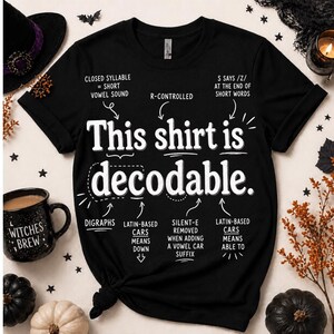 Op de afbeelding: Een zwart T-shirt met korte mouwen en witte tekst: This shirt is decodable. Rondom de tekst staan taalkundige aantekeningen: Closed syllable = short vowel sound, R-controlled, S says /z/ at the end of short words, Digraphs, Latin-based cars means down, Silent-e removed when adding a vowel car suffix en Latin-based cars means able to.