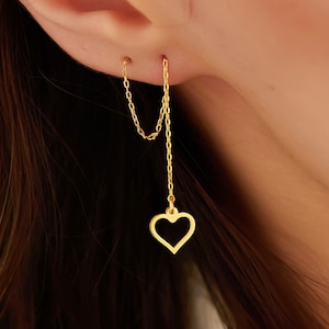 May include: Gold-colored heart-shaped earrings. The earrings feature a small heart pendant suspended from a delicate chain. The chain connects to a small stud that goes through the ear. The earrings are a simple and elegant design.