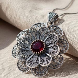 Op de afbeelding: Een zilveren hanger aan een ketting met een meerlaags bloemmotief gemaakt van ingewikkeld filigraan draadwerk. Het midden van de bloemvormige hanger is bezet met een ronde, gefacetteerde dieprode edelsteen, hangend aan een fijne zilveren ketting.