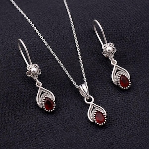 May include: A silver jewellery set comprising a necklace and earrings. Each piece features a teardrop-shaped, dark red gemstone. The earrings have a floral design at the top. The necklace has a matching pendant.