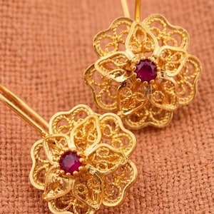 May include: A pair of gold earrings featuring a floral filigree design with intricate wirework patterns. Each earring is centered with a small, round, deep red gemstone. The earrings are attached to thin, curved gold ear wires for hanging.