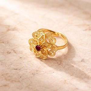 May include: A gold-colored ring featuring a filigree flower design with a small, round, red gemstone at the center. The ring is set against a light-colored, marble-like surface.