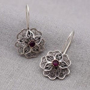 Puede incluir: Un par de pendientes colgantes de tono plateado con un diseño floral de filigrana. Cada pendiente tiene una piedra circular de color rojo oscuro en el centro, rodeada de intrincados pétalos de alambre metálico y un gancho curvo.