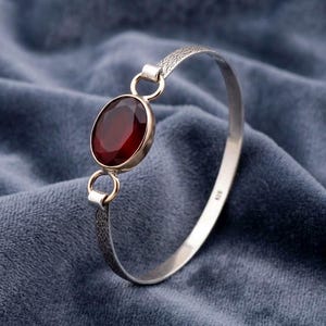 May include: Silver bangle bracelet with a large, oval, deep red gemstone. The gemstone is set in a gold-toned bezel. The bracelet has a textured design on the connecting links and a smooth, polished band.