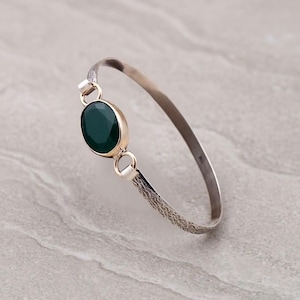 May include: A silver bangle bracelet featuring a gold-toned setting with a large, oval, dark green gemstone. The bracelet has a textured design on the ends and a simple, curved band.