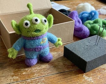 May include: A needle-felted green alien figure with three eyes, pointed ears, and a single antenna, wearing a blue suit with a purple belt and a yellow planet emblem. The figure is positioned next to a cardboard box, wool roving, and a foam block with needles.