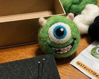 May include: A needle felting kit featuring a round green monster character with one large blue eye, small horns, and a wide smile. The kit includes a cardboard box, loose green, white, and black wool roving, a black foam block, needles, and an instruction sheet.