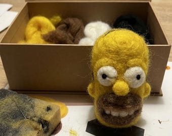 May include: A needle felted yellow character head with white eyes, a brown beard, and white teeth. The kit includes a cardboard box containing yellow, brown, white, and black wool roving, a foam block, and an instruction sheet with the text INSTRUCTIONS.