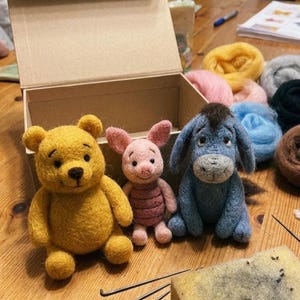 May include: Three needle-felted figures of a yellow bear, a small pink pig, and a blue donkey with a dark mane. The figures are positioned in front of an open cardboard box, with colorful wool roving bundles and felting needles nearby.