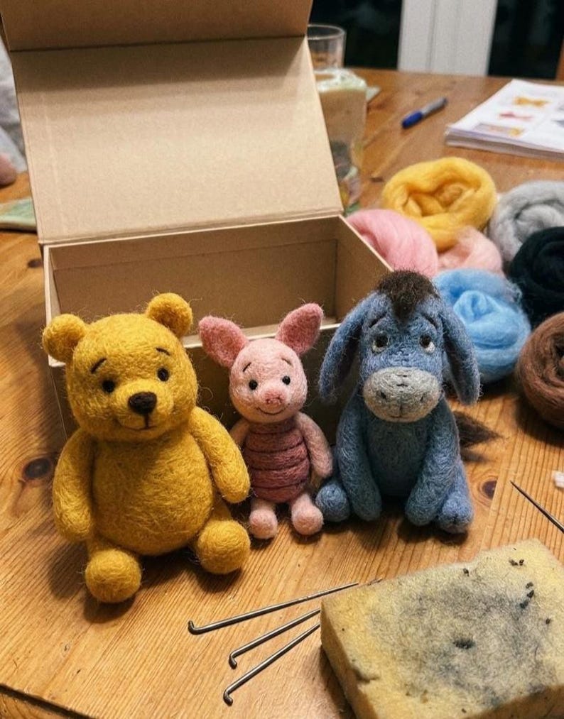 May include: Three needle-felted figures of a yellow bear, a small pink pig, and a blue donkey with a dark mane. The figures are positioned in front of an open cardboard box, with colorful wool roving bundles and felting needles nearby.